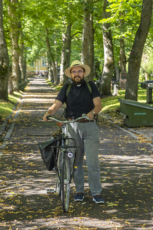 Pappi, tumma pusero ja hellehattu sekä polkupyörä. Hautausmaan kaunis vehreä käytävä.