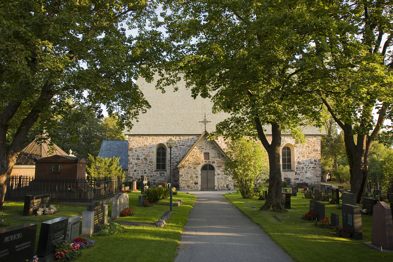 Stone church building. In front of it a graveyard and lots of green leaved trees. Photo Timo Jakonen.