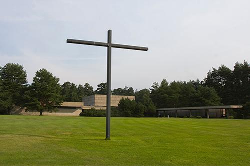 Big cross on a field. Behind it a low building and trees.