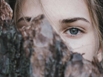 Nuoren naisen silmä näkyy puun kuoren takaa, luoden salaperäisen tunnelman. Hänen katseensa on intensiivinen ja herkkä.