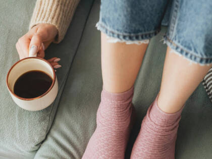 En person sitter på en soffa med en kaffekopp bredvid sig. Man ser bara fötterna på personen.