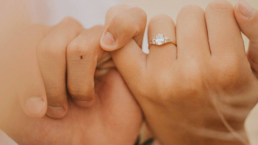 Two hands holding each other by the pinkie fingers. One hand has a diamond ring.