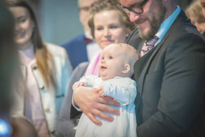 Vauva hymyilee isänsä sylissä. Ympärillä seisoo hymyileviä ihmisiä.