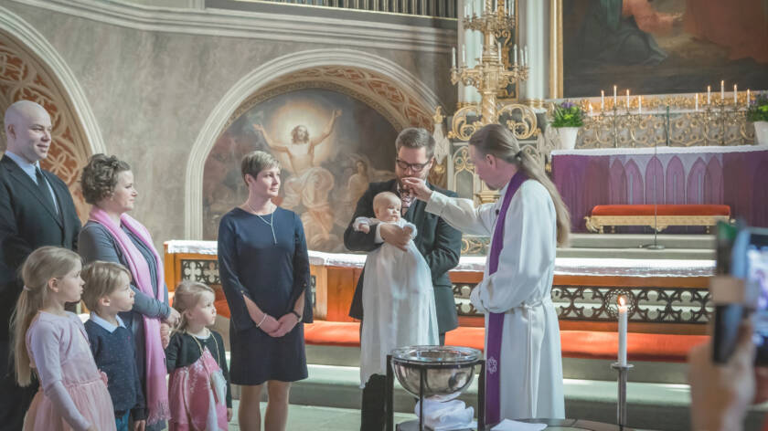 Pappi tekee ristinmerkin vauvan pään päälle. Ympärillä on ihmisiä katsomassa.