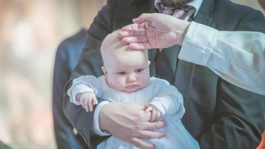 En baby i vit klänning hålls i sin pappas famn. Pappan rör vid babyns huvud, en suddig kyrka i bakgrunden.