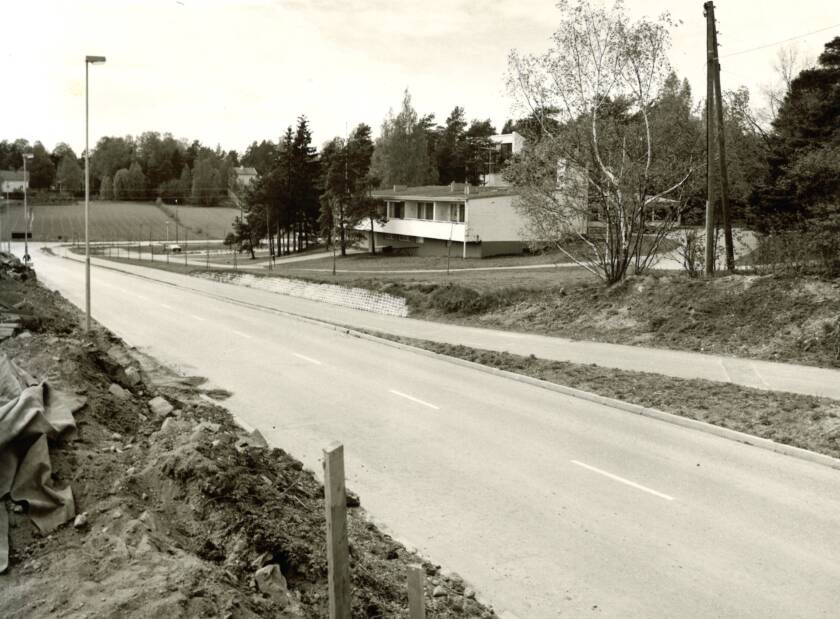 Mustavalkoinen kuva, jossa näkyy tyhjää tietä, puut ja matala rakennus taustalla. Tie laskee alas kohti rakennusta.
