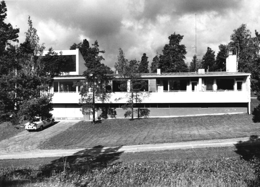 Moderni rakennus metsäisessä ympäristössä, jossa on suuri terassi ja useita ikkunoita. Taustalla näkyy puita ja pilvinen taivas.