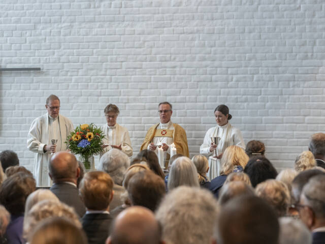 Neljä pappia seisoo alttarilla, taustalla valkoinen tiiliseinä. Edessä on kukkakimppu ja seurakunta kuuntelee.