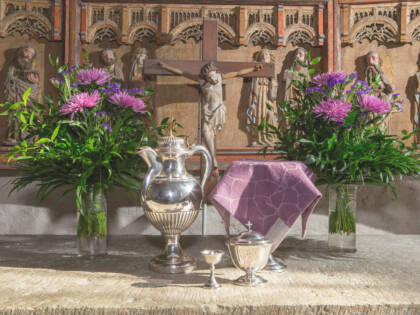 Ehtoollismalja, viinikannu, ehtoollisleipien astia ja pieni pikari aseteltuna keskiaikaisen kirkon alttarille, jonka keskellä on krusifiksi. Alttarilla olevat kukat ja ehtoollismaljaa peittävä liina ovat liilat.