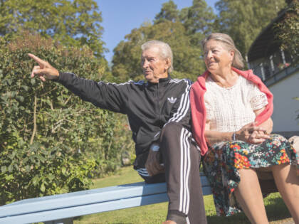 Eläkeläispariskunta istuu penkillä. Mies osoittaa kuvan ulkopuolelle.
