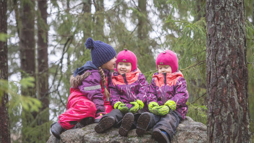 Kolme pikkutyttöä istuu kivellä metsässä.
