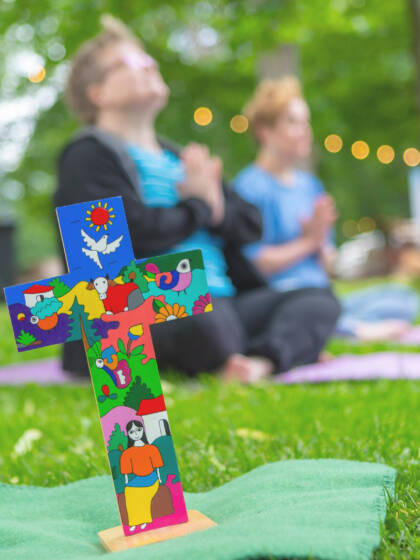 Värikäs risti, jonka takana kaksi naista istuu jalat ristissä meditoimassa.