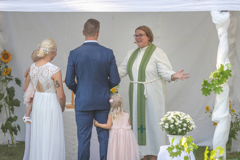 Prästen tar emot brudparet med utsträckta armar.