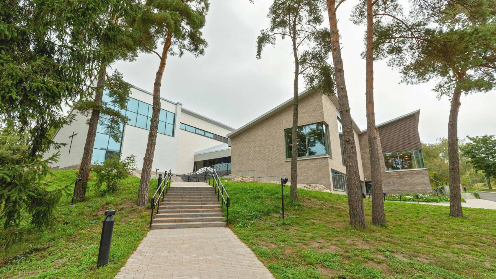 Vaalea, moderni tiilirakennus mäen päällä. Edustalla rakennukselle johtavat portaat ja mäntyjä.