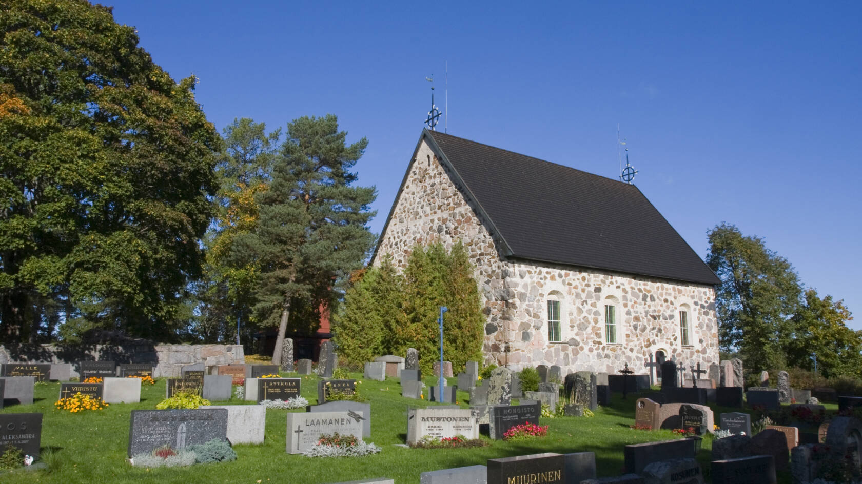 En gammal stenkyrka i mitten av en gravgård, omgiven av gröna träd och en klarblå himmel. Gravstenar är ordnade på gräset.