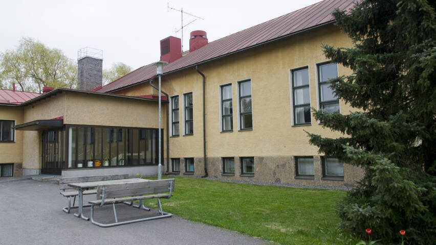 Keltainen, rapattu rakennus, jossa ruutuikkunat. Edustalla pöytä ja kaksi penkkiä.