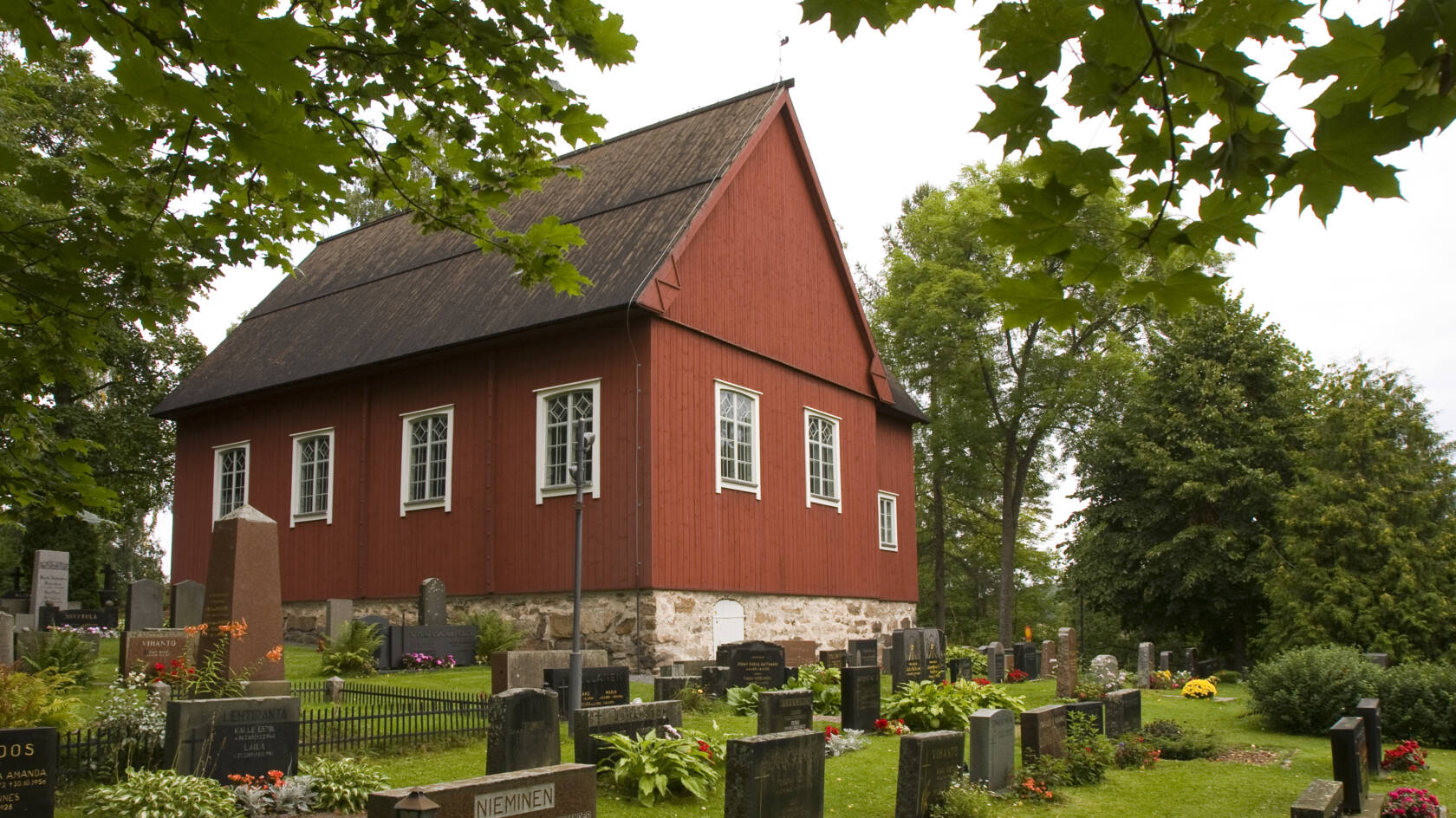 Röd kyrka vid kanten av en kyrkogård, omgiven av gröna träd och blommor. Gravstenar i olika former och färger.