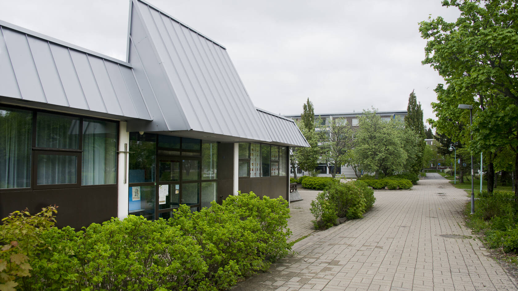 Moderni, matala rakennus, jossa lasiovet. Edustalla laatoitettu kulkureitti ja pensasrivi.