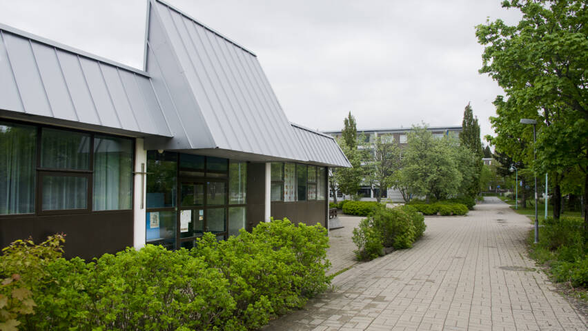 Moderni, matala rakennus, jossa lasiovet. Edustalla laatoitettu kulkureitti ja pensasrivi.