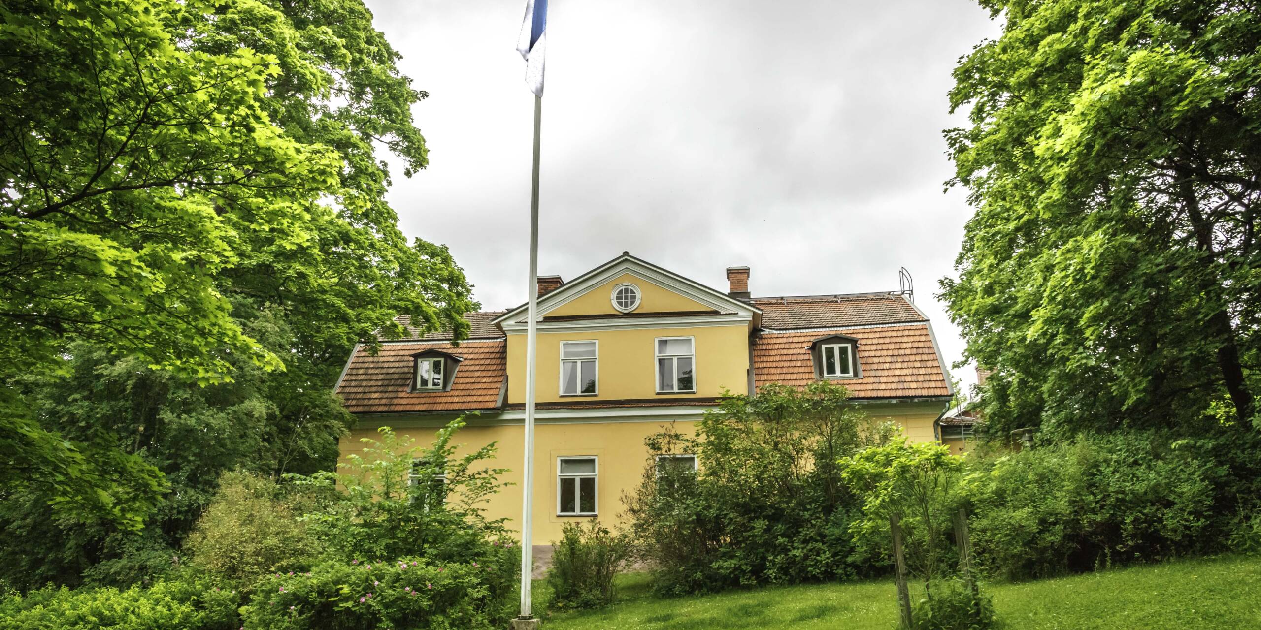 Ett gammalt gult hus omgiven av gröna träd, med den finska flaggan hissad, i bakgrunden grå moln.