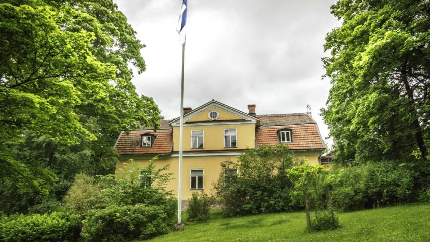 Vanha keltainen talo vihreiden puiden ympäröimänä, Suomen lippu salkoon nostettuna, taustalla harmaita pilviä.