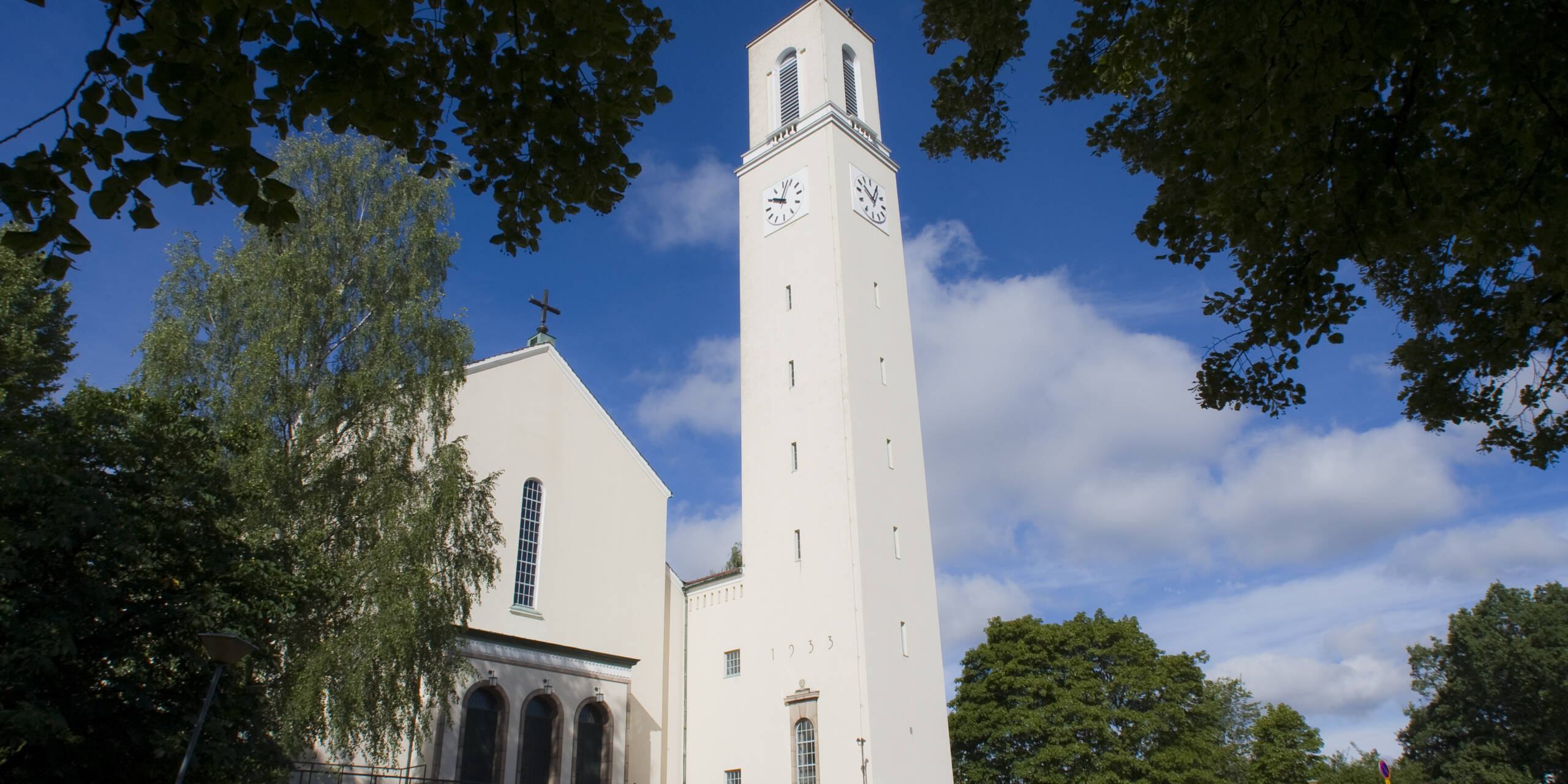 Valkoinen kirkko, jossa on korkea kellotorni ja suuret kellot, ympäröity vihreillä puilla ja sinisellä taivaalla.