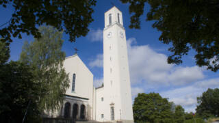 En vit kyrka med ett högt klocktorn och stora klockor, omgiven av gröna träd och en blå himmel.