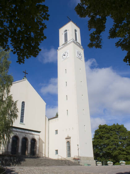 Valkoinen kirkko, jossa on korkea kellotorni ja suuret kellot, ympäröity vihreillä puilla ja sinisellä taivaalla.