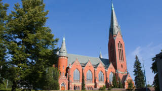 Vacker tegelkyrka med ett högt torn och intrikata fönster, omgiven av gröna träd och en blå himmel.