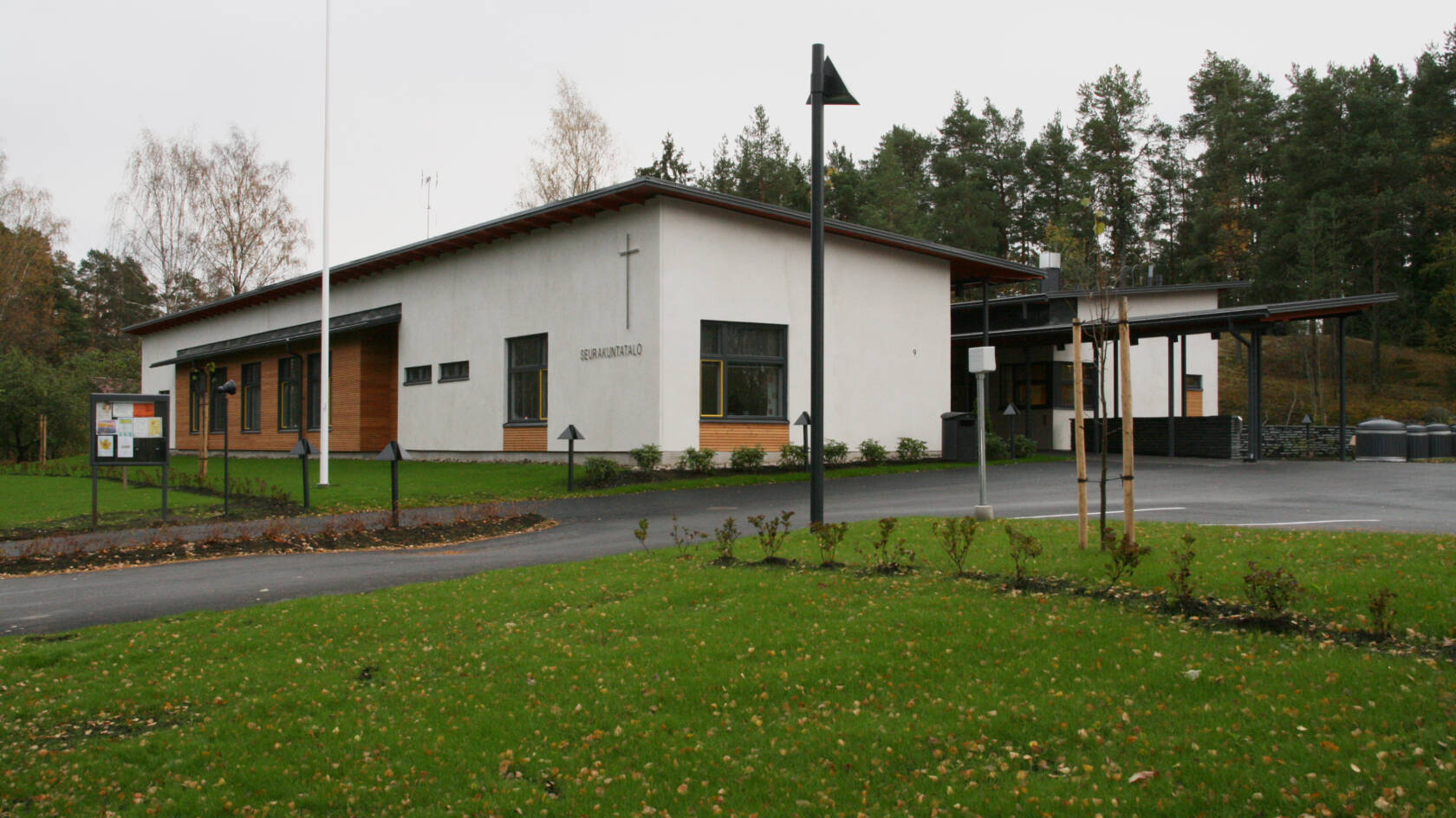 Moderni rakennus, jossa on valkoinen julkisivu ja risti. Edessä nurmikkoa ja puita, taustalla metsää.