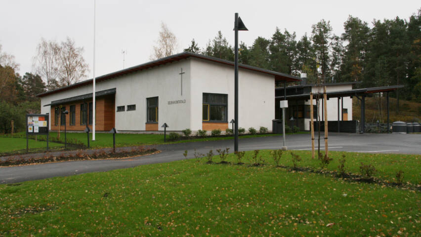Moderni rakennus, jossa on valkoinen julkisivu ja risti. Edessä nurmikkoa ja puita, taustalla metsää.