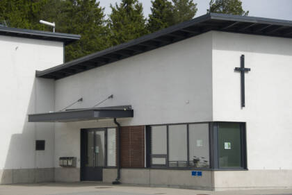Valkoinen, mustakattoinen moderni rakennus. Ulko-oven päällä lippa ja ulkoseinällä risti.