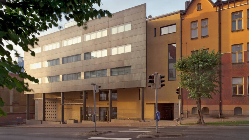 Modern byggnad med glasfasad och en gul tegelbyggnad bredvid. Ett övergångsställe och träd syns i förgrunden.