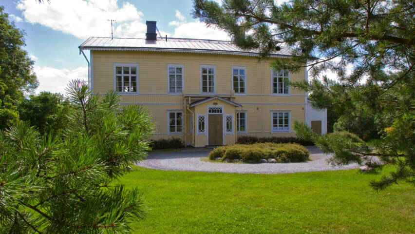 Kaunis keltainen talo, jossa on valkoiset ikkunat ja ovi, ympäröity vihreällä nurmikolla ja puilla. Taustalla kirkas taivas.