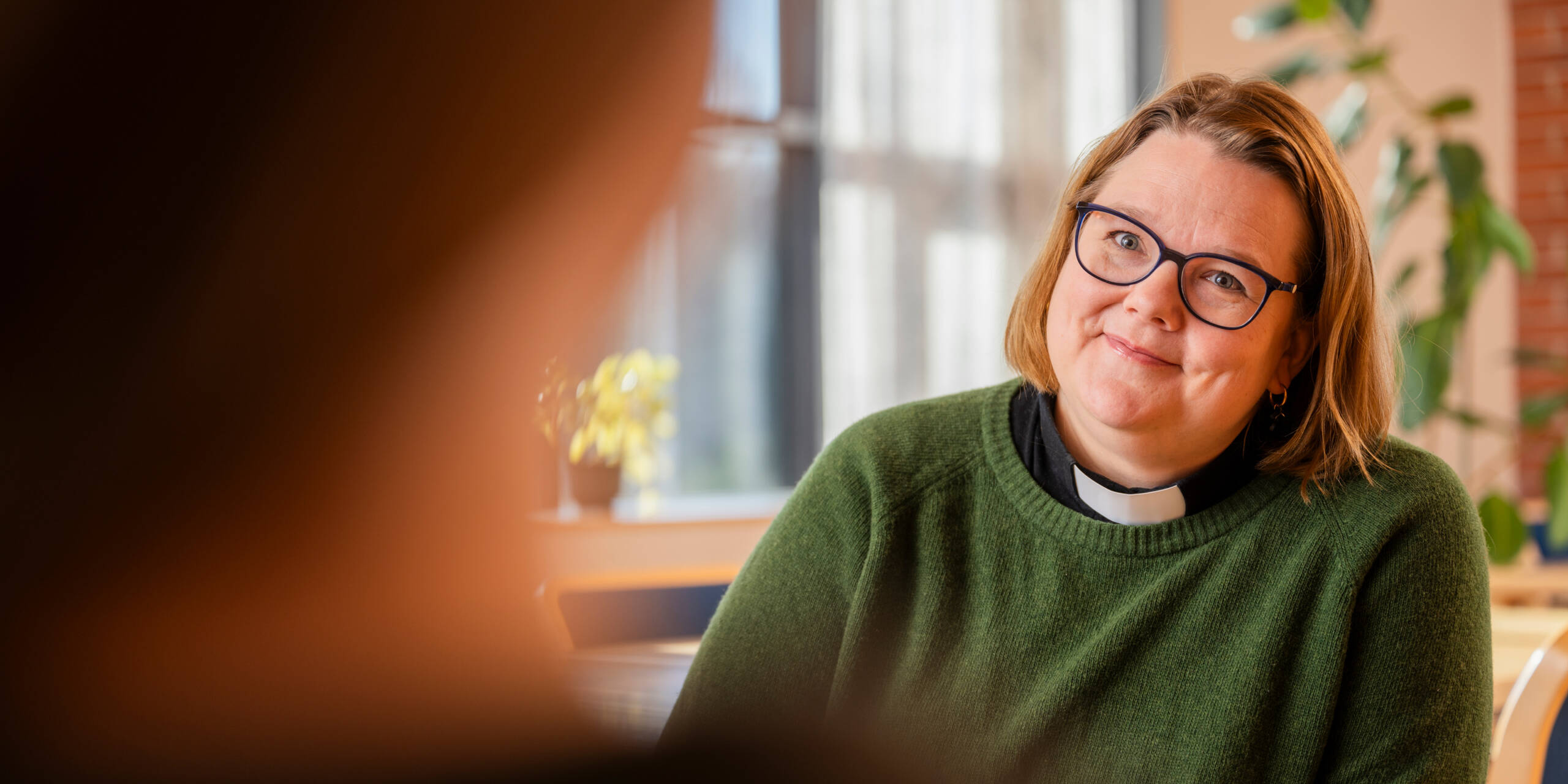 Vihreään neuleeseen pukeutunut pappi katsoo hymyillen häntä vastapäätä istuvaa henkilöä. Taustalla kasveja ja ikkuna, valo tulvii huoneeseen.