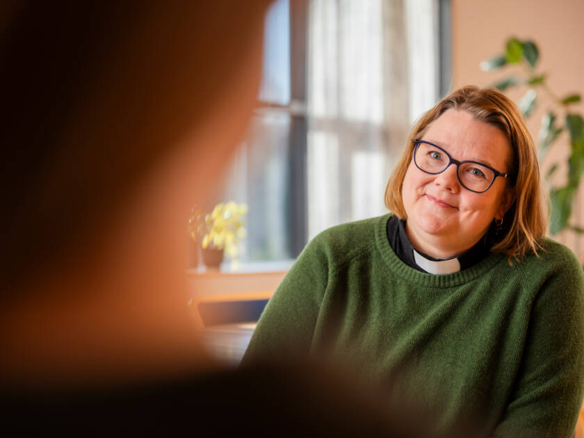 Vihreään neuleeseen pukeutunut pappi katsoo hymyillen häntä vastapäätä istuvaa henkilöä. Taustalla kasveja ja ikkuna, valo tulvii huoneeseen.