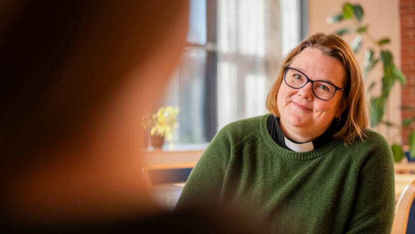 Vihreään neuleeseen pukeutunut pappi katsoo hymyillen häntä vastapäätä istuvaa henkilöä. Taustalla kasveja ja ikkuna, valo tulvii huoneeseen.
