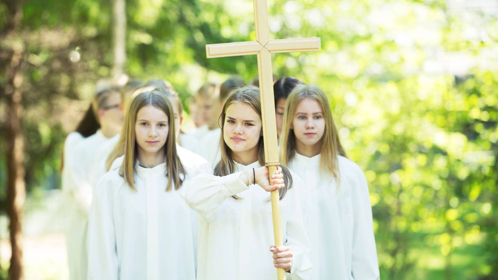 Nuoria kulkee valkoisissa alboissa parijonossa. Ensimmäisenä on ristiä kantava tyttö.