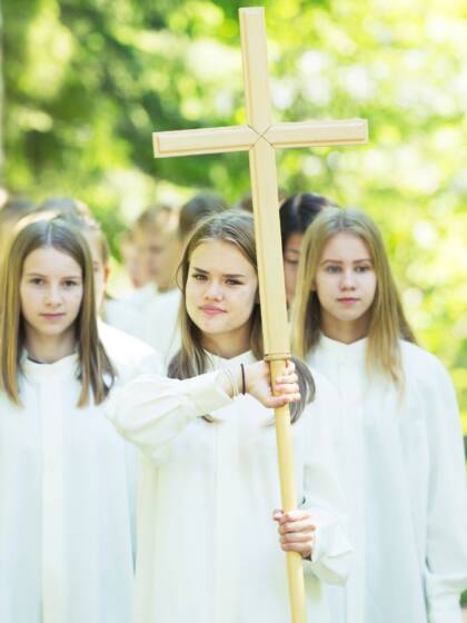 Nuoria kulkee valkoisissa alboissa parijonossa. Ensimmäisenä on ristiä kantava tyttö.