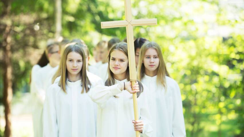 Nuoria kulkee valkoisissa alboissa parijonossa. Ensimmäisenä on ristiä kantava tyttö.