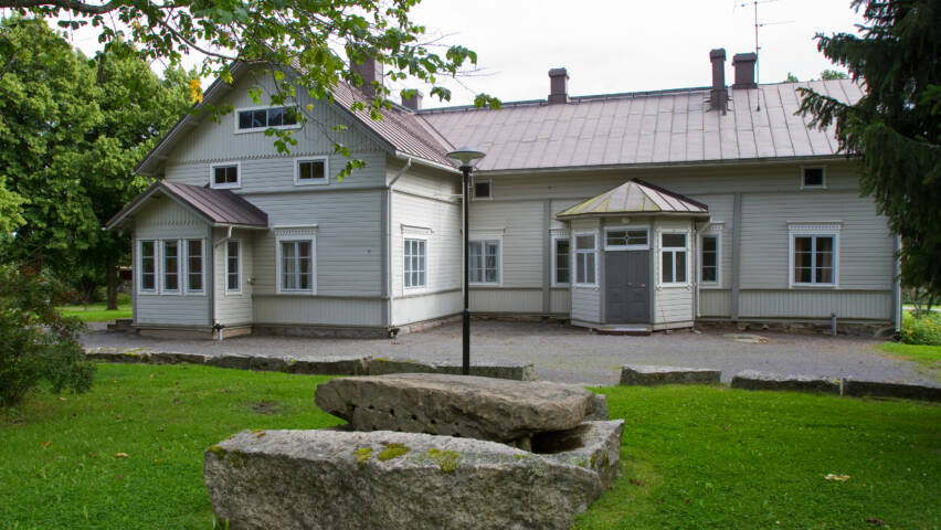 Vanha vaaleanharmaa puutalo, jossa on valkoiset ikkunat ja harmaa ovi. Edessä kiviä ja vihreää nurmikkoa. Taustalla puita ja kirkas taivas.