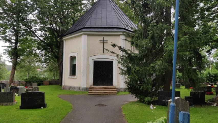 Pyöreä, vaaleankeltainen siunauskappeli, jossa on musta katto ja risti. Ympärillä vihreää nurmikkoa ja puita, hautakiviä näkyvissä.