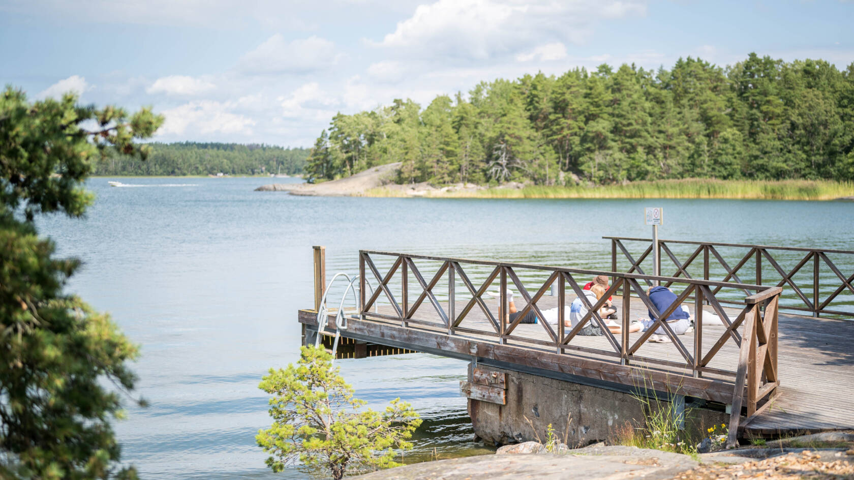 Kaunis laituri meren rannalla, jossa on vihreitä puita taustalla. Aurinko paistaa ja ihmiset nauttivat kesäpäivästä.