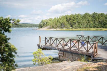 Kaunis laituri meren rannalla, jossa on vihreitä puita taustalla. Aurinko paistaa ja ihmiset nauttivat kesäpäivästä.