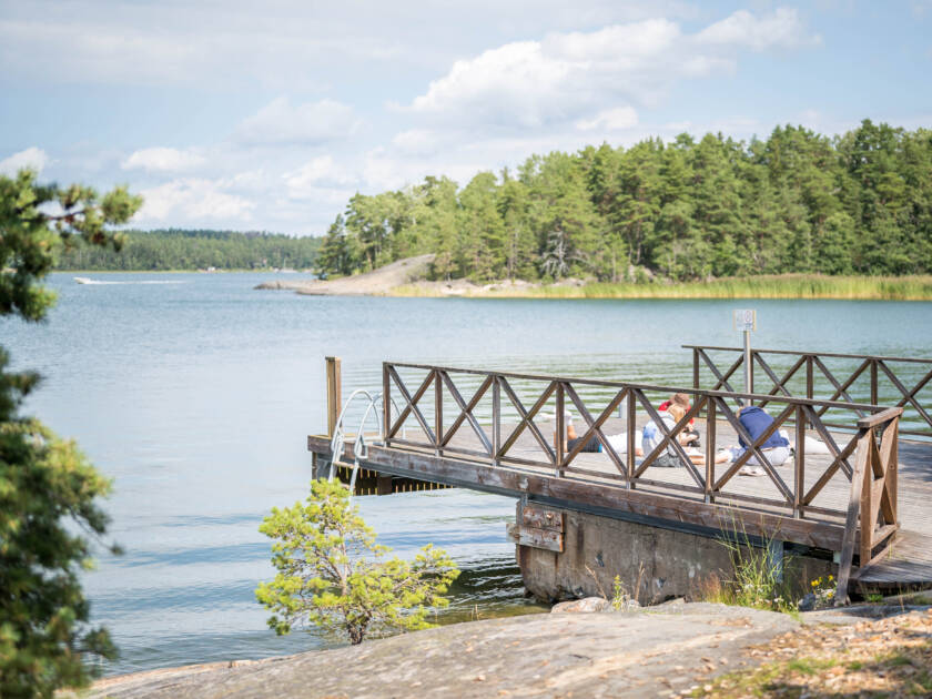 Kaunis laituri meren rannalla, jossa on vihreitä puita taustalla. Aurinko paistaa ja ihmiset nauttivat kesäpäivästä.