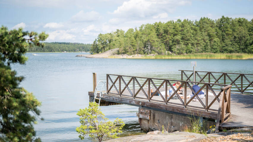 Kaunis laituri meren rannalla, jossa on vihreitä puita taustalla. Aurinko paistaa ja ihmiset nauttivat kesäpäivästä.