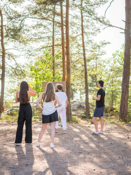 Neljä nuorta seisoo selin kameraan, metsän keskellä. Taustalla näkyy järvi ja auringonvalo suodattuu puiden läpi.
