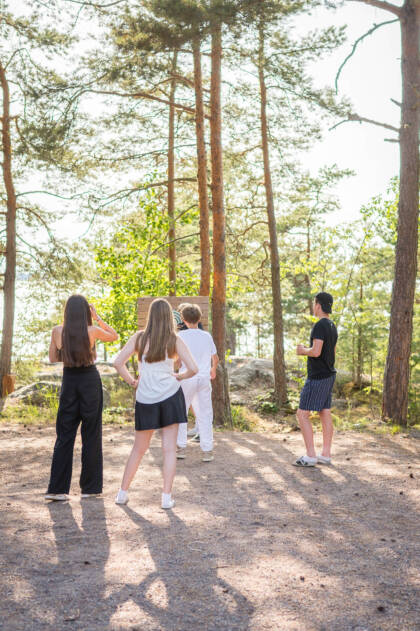 Neljä nuorta seisoo selin kameraan, metsän keskellä. Taustalla näkyy järvi ja auringonvalo suodattuu puiden läpi.
