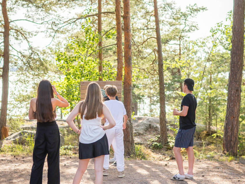 Neljä nuorta seisoo selin kameraan, metsän keskellä. Taustalla näkyy järvi ja auringonvalo suodattuu puiden läpi.