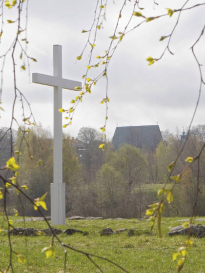 Valkoinen risti keskellä peltoa. Taustalla näkyy vanha kivikirkko.